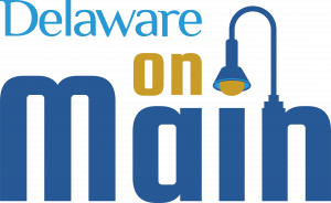 Logo of Delaware on Main program. Features Delaware in smaller light blue letters, the word on, in gold letters - and that sits over the word Main with a capital M. A lampost sprouts out of the top of n in Main, and it curves over to end in a liamplight on top of the letter i in Main. 